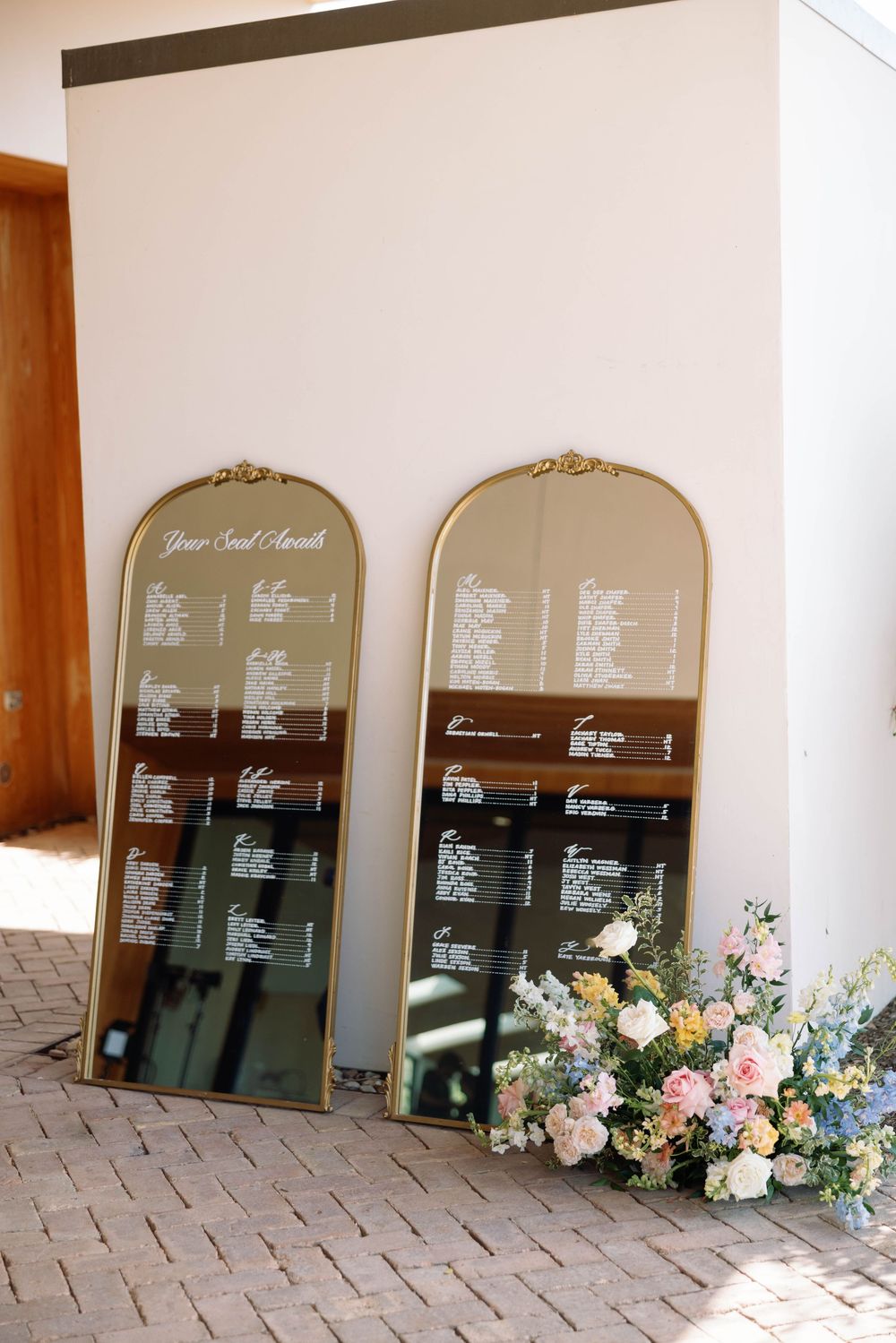 Two arched seating chart mirrors decorated with elegant floral arrangements in pink and blue sit on a stone courtyard.