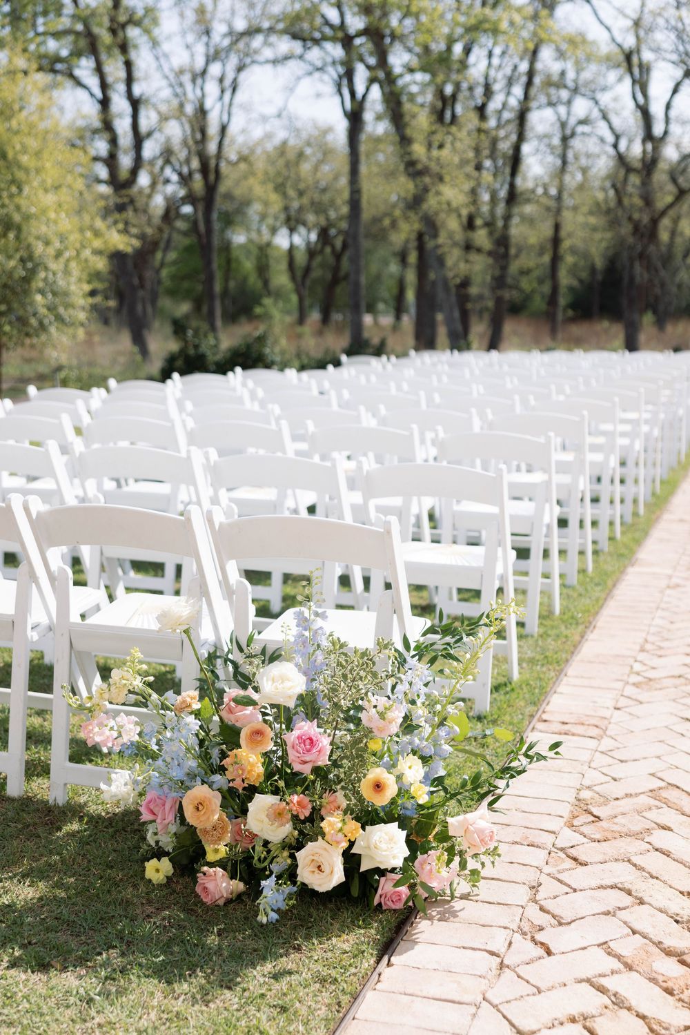 Rows of white folding chairs along a brick pathway with a lush floral arrangement of pink and peach roses for outdoor wedding.