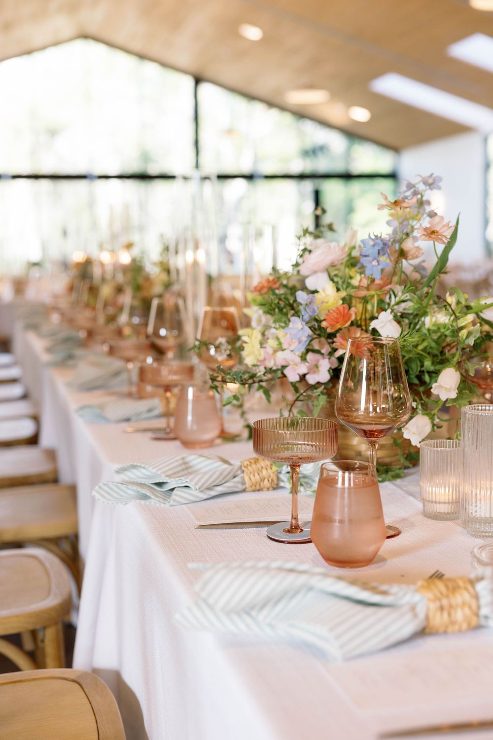Elegant wedding table setting with floral centerpieces, blush glassware and striped napkins under a tent-style venue.