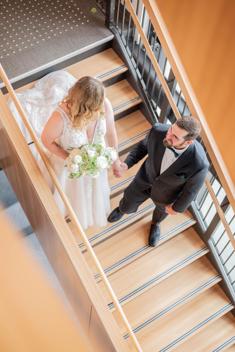 Wedding couple walks down wide modern staircase, captured from above showing bride's dress and groom's suit.