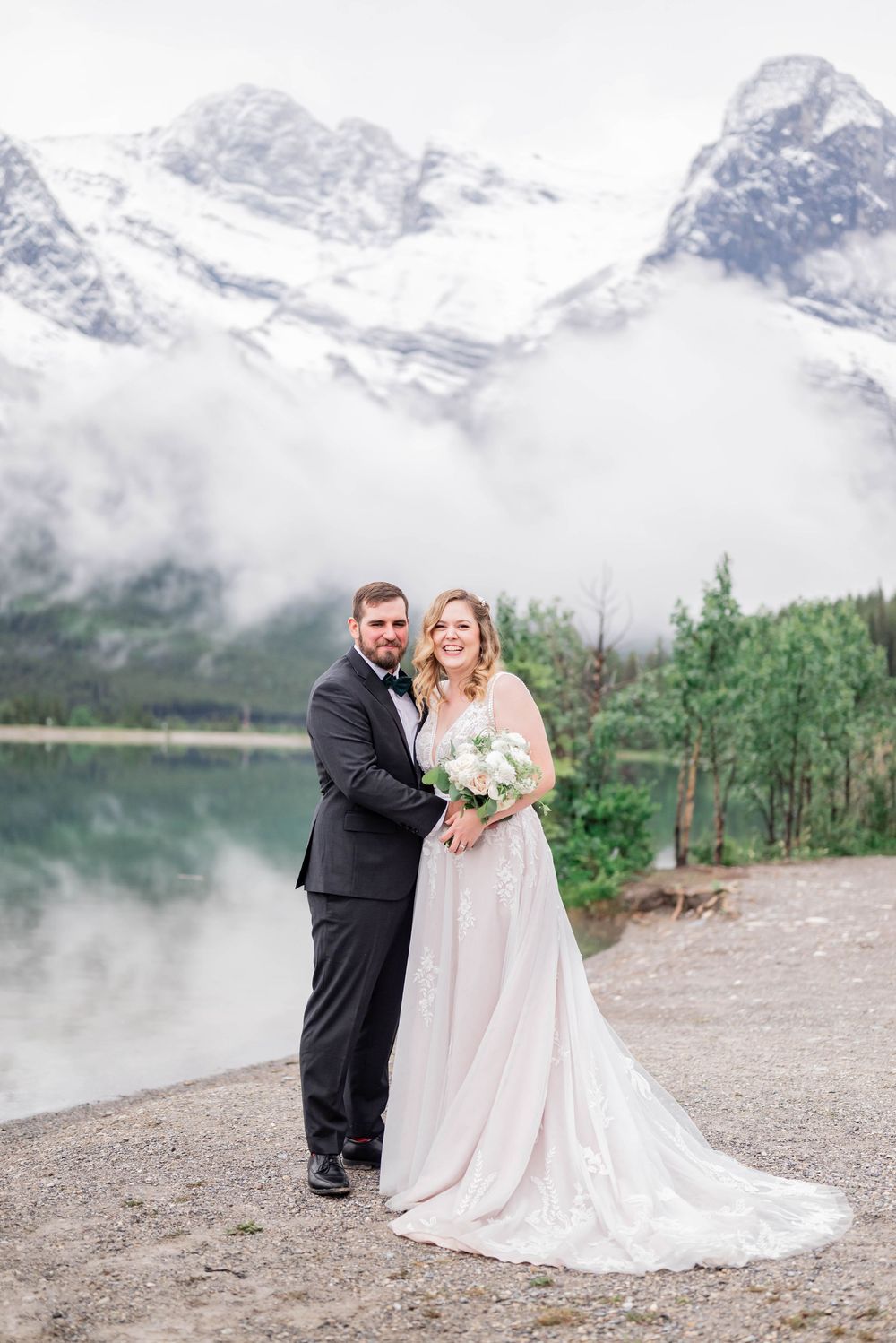 A wedding couple poses by a misty mountain lake with snow-capped peaks and evergreen trees in the background.