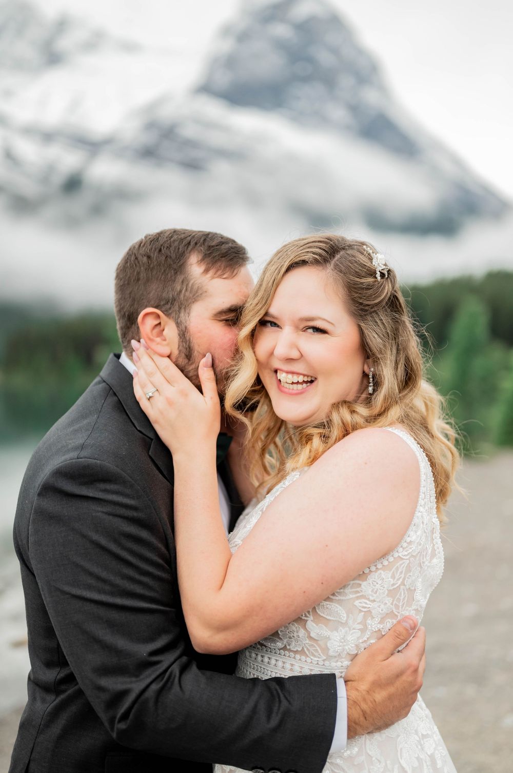 A romantic wedding couple embraces and laughs together with a dramatic mountain backdrop in the clouds.