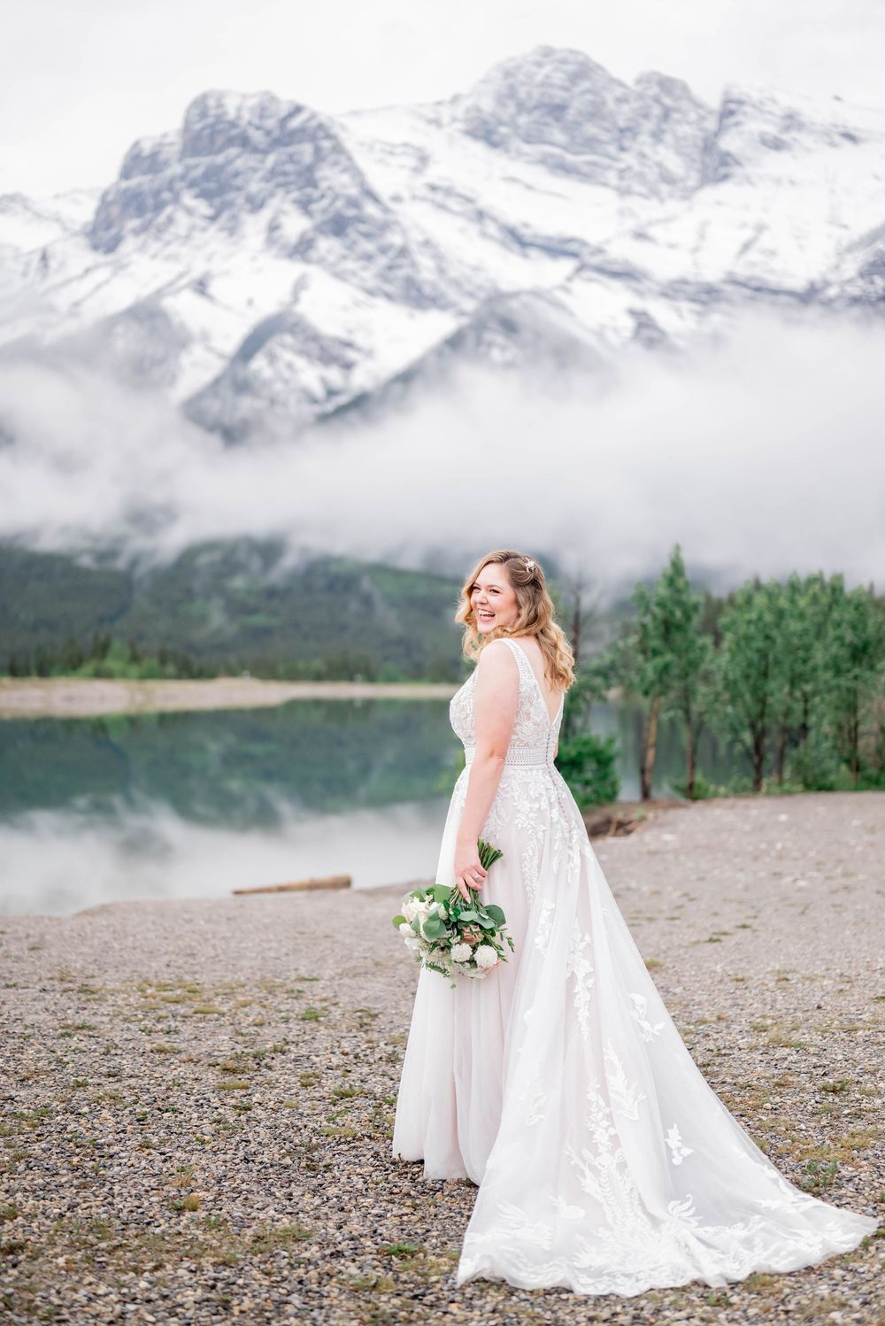 A bride in a flowing white gown holds a bouquet against a backdrop of misty mountains and an alpine lake.