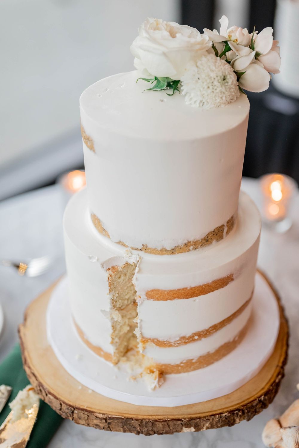 Three-tier white wedding cake with exposed layers and delicate floral decorations on a rustic wood slice base.