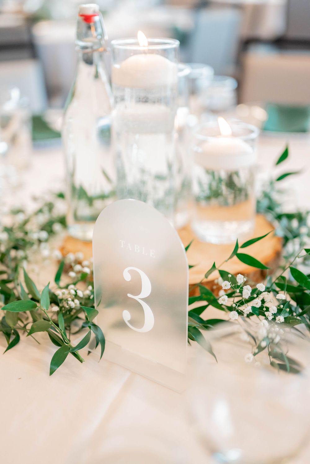 Elegant wedding table setting with frosted glass table number 3 surrounded by greenery and glowing candles.