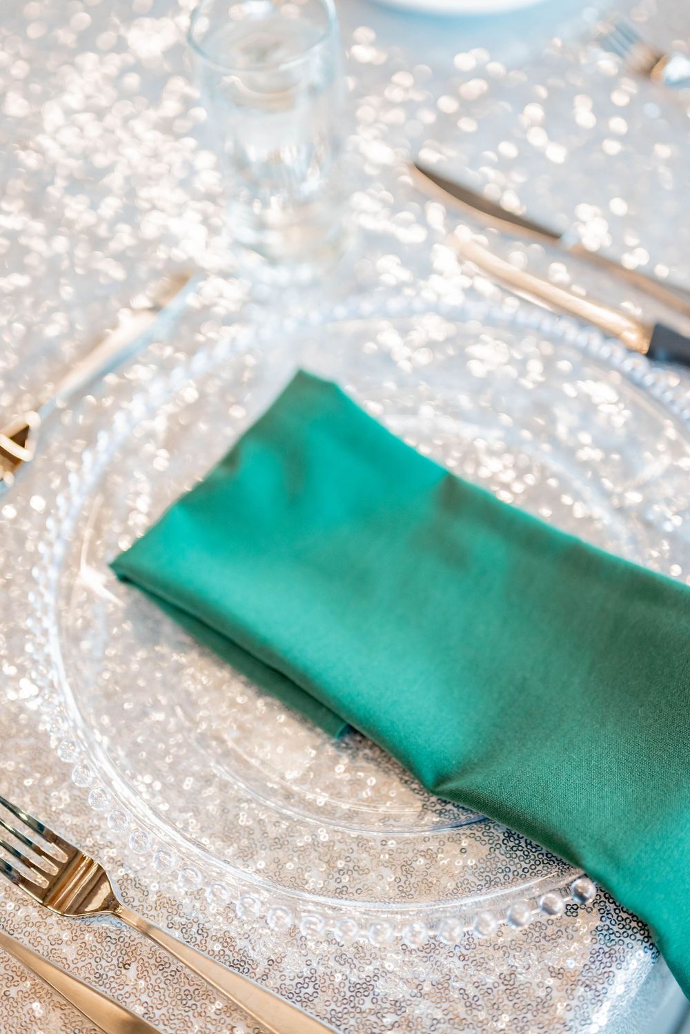 Clear glass charger plate with turquoise napkin and gold utensils on sparkly festive table setting.