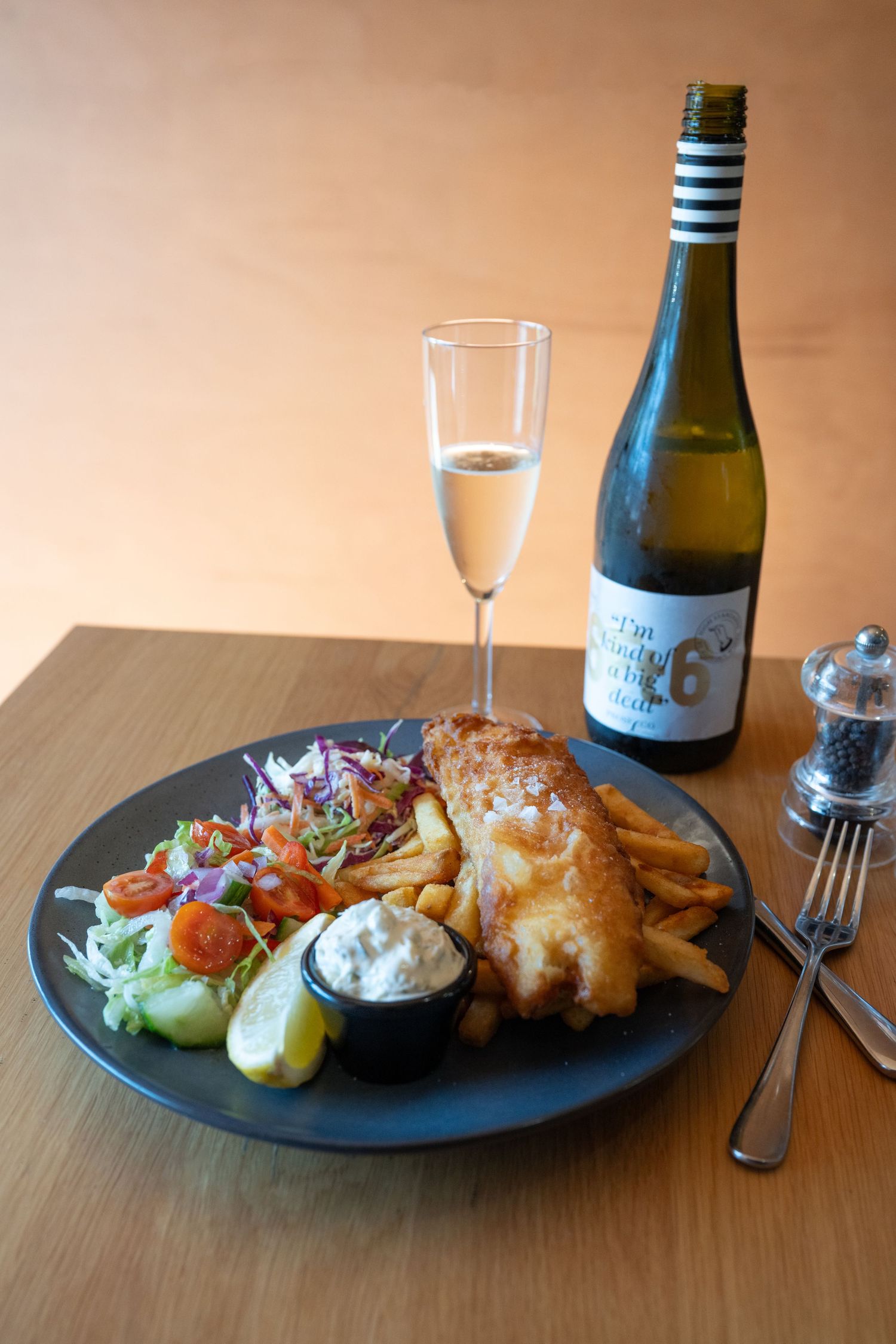 Fish and chips dinner plate served with fresh salad, tartar sauce and white wine on wooden table.