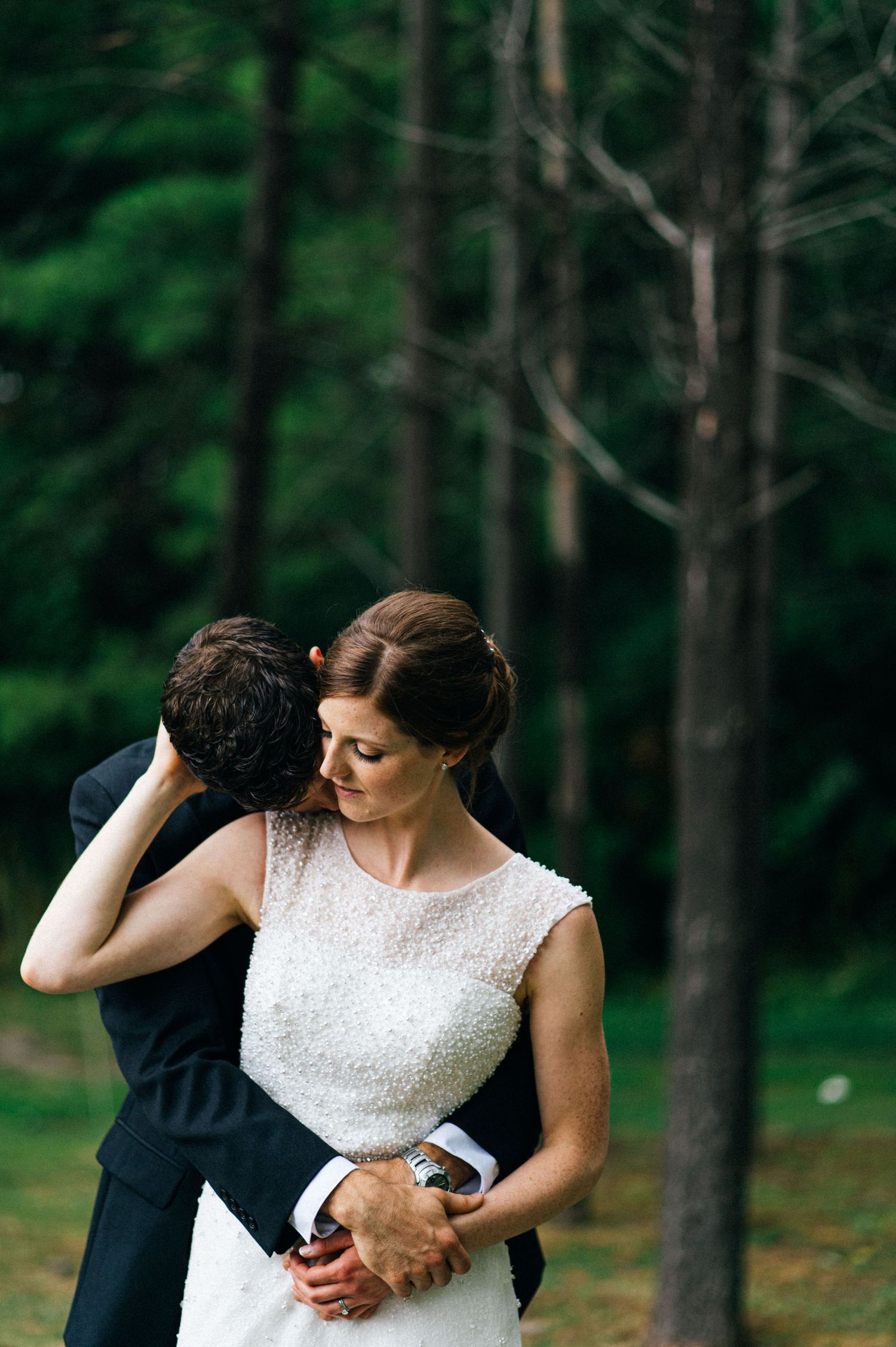 A tender wedding moment captured between a couple in a forest setting with the bride wearing a sleeveless white dress.