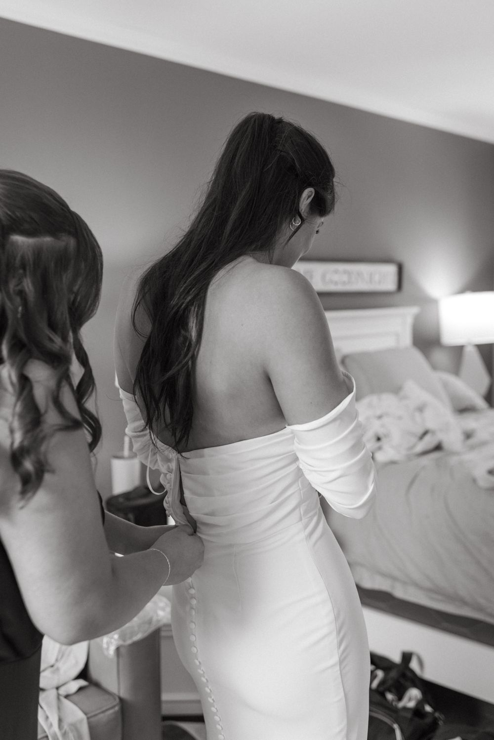 A woman in an off-shoulder white dress stands in a bedroom as she prepares for a special occasion.