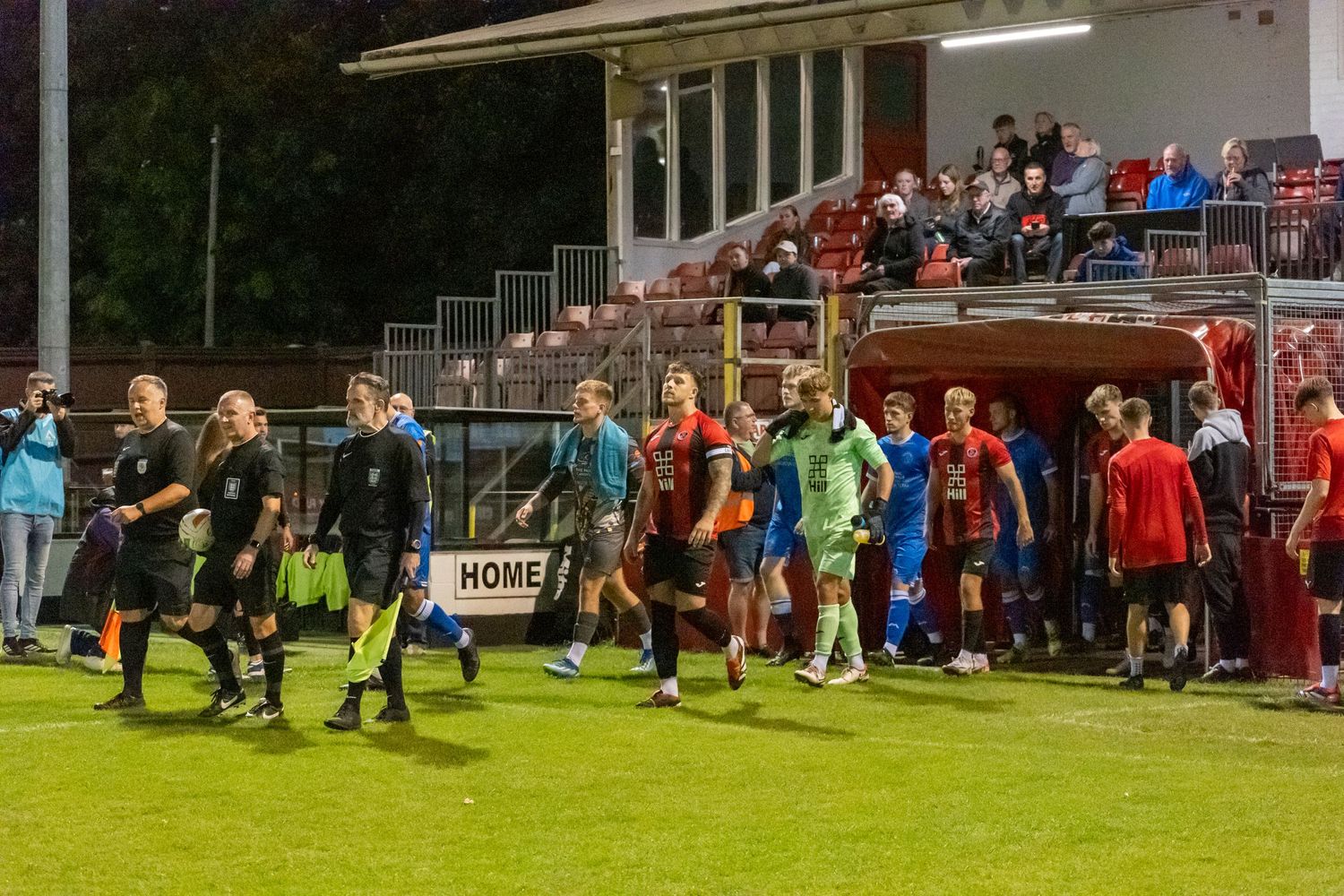 Soccer teams walk onto the field before a night match with spectators watching from stadium stands in the background.