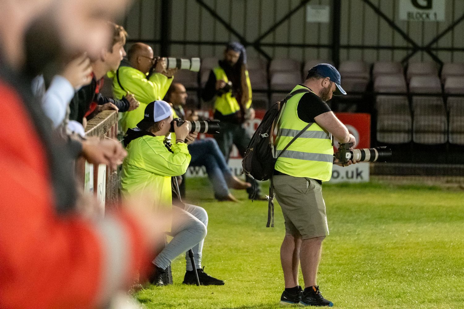 Sports event stewards in high-visibility vests monitoring activity from sidelines of a grass playing field during match.