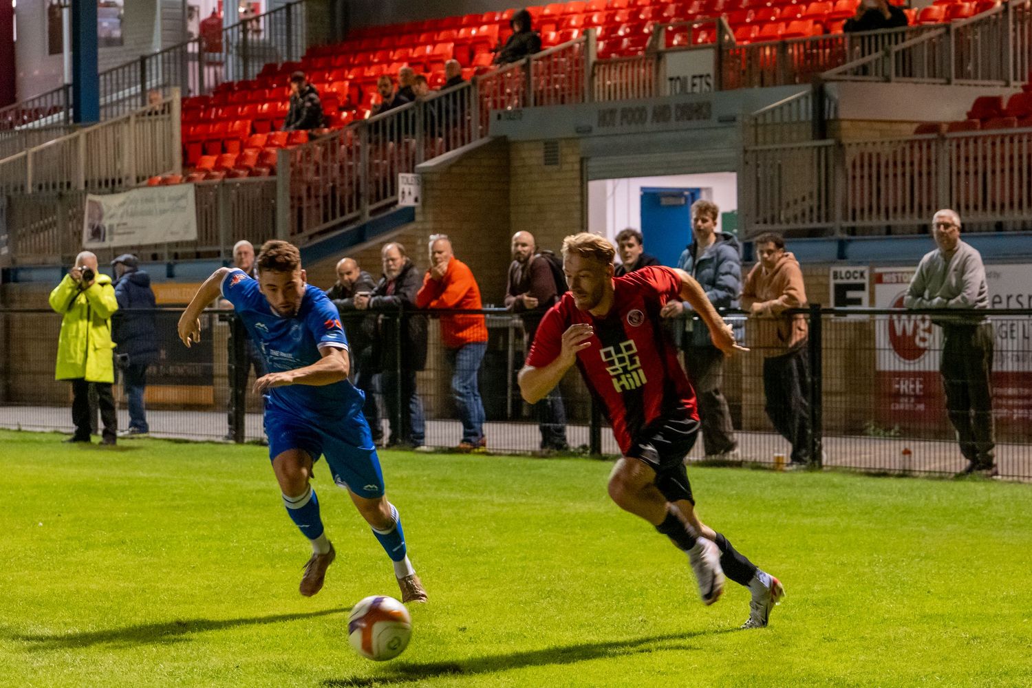 Two soccer players compete for the ball during a night match under red stadium lights.
