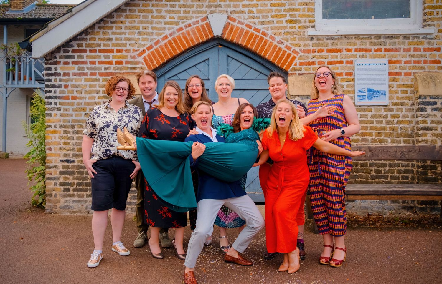 Fun group wedding portrait with friends lifting someone in celebration outside a brick building.