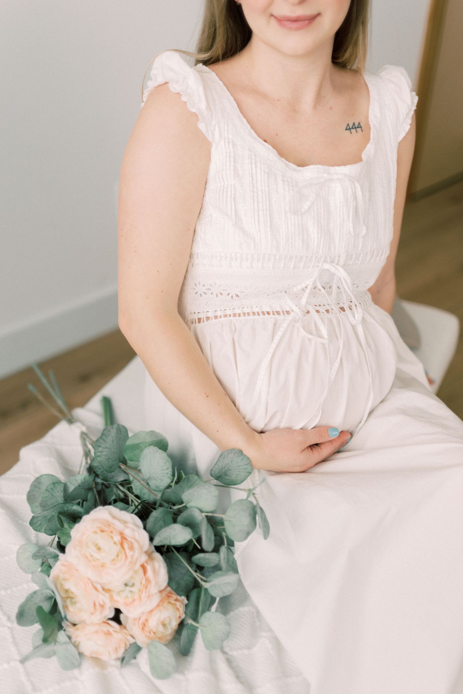 White maternity dress with lace details and peach roses with eucalyptus bouquet in a soft, feminine photo setting.
