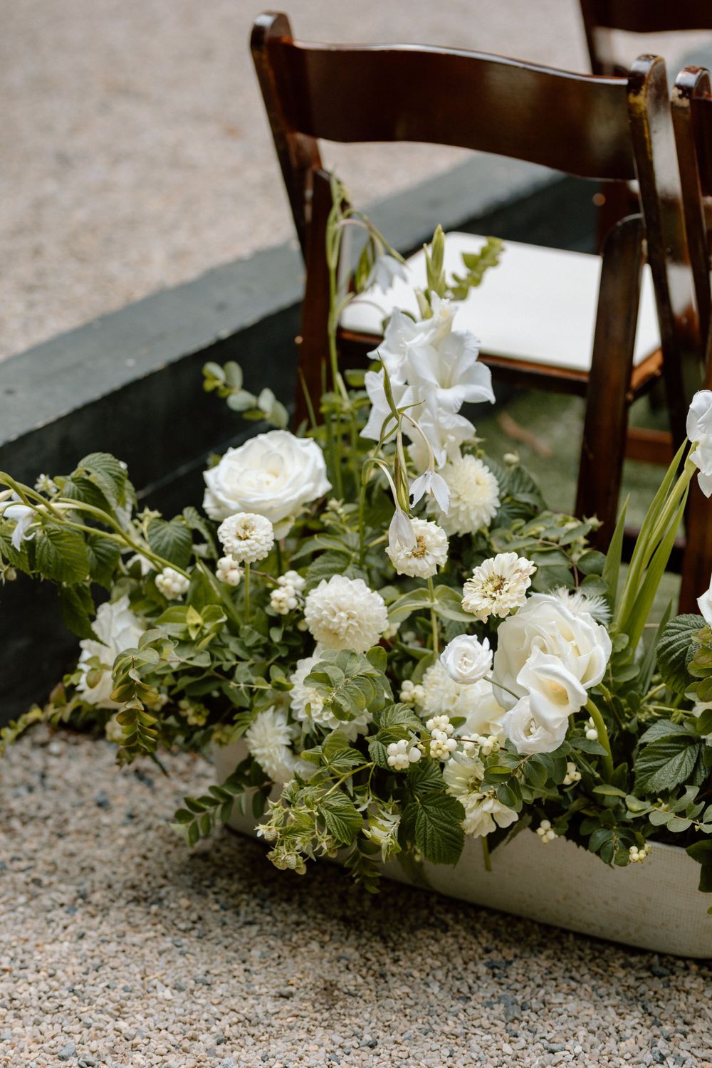 A rustic wooden chair beside an elegant floral arrangement with white roses and greenery on a concrete floor.