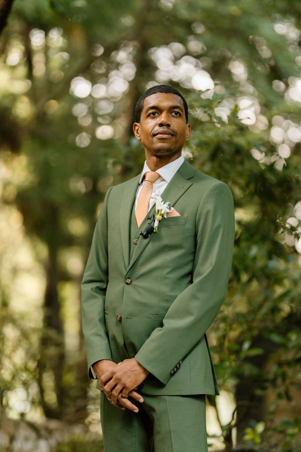 Well-dressed groom in sage green suit with peach boutonniere poses outdoors against natural bokeh background in forest.