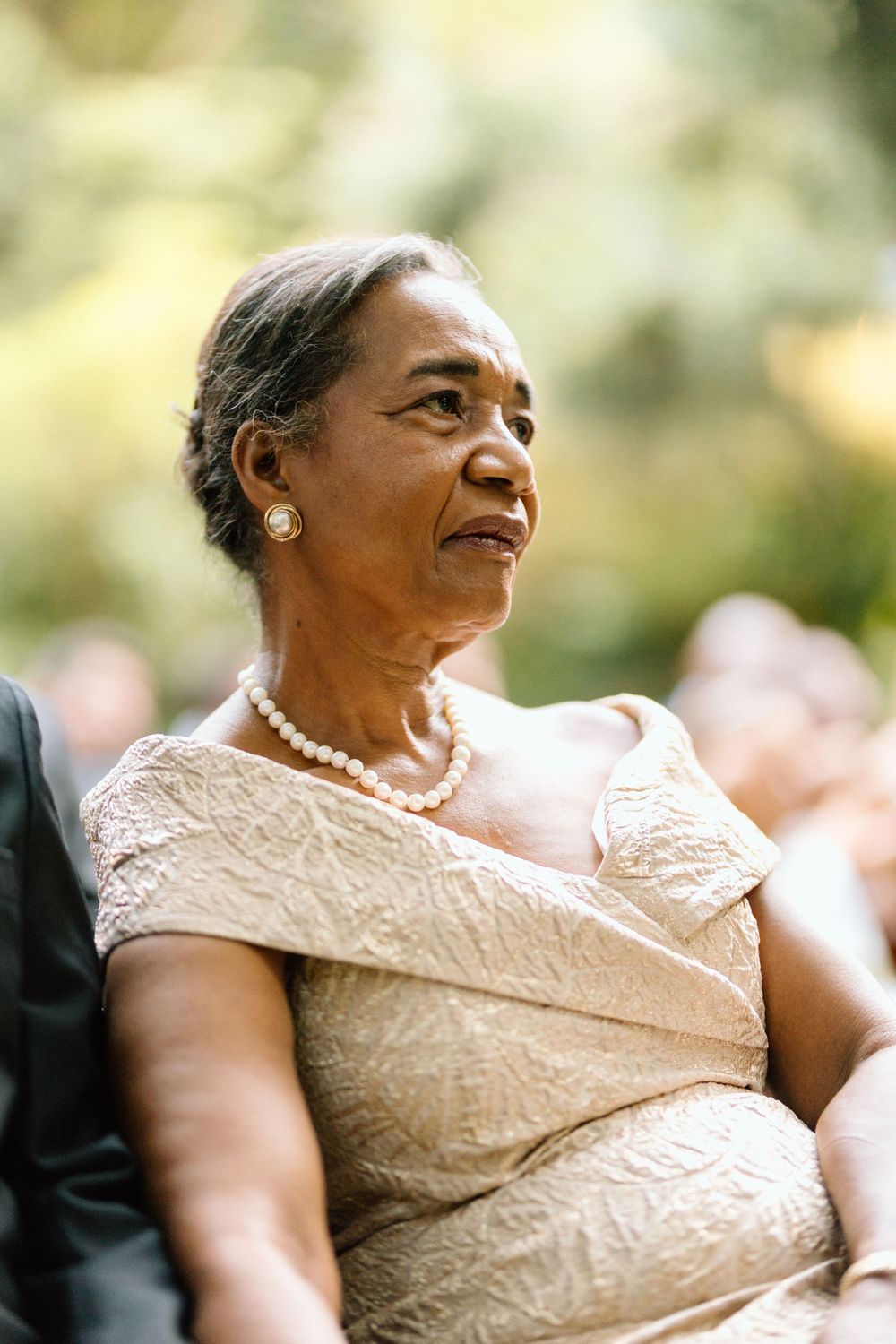 An elegant woman in a cream-colored lace dress and pearl necklace sits gracefully at a formal occasion.