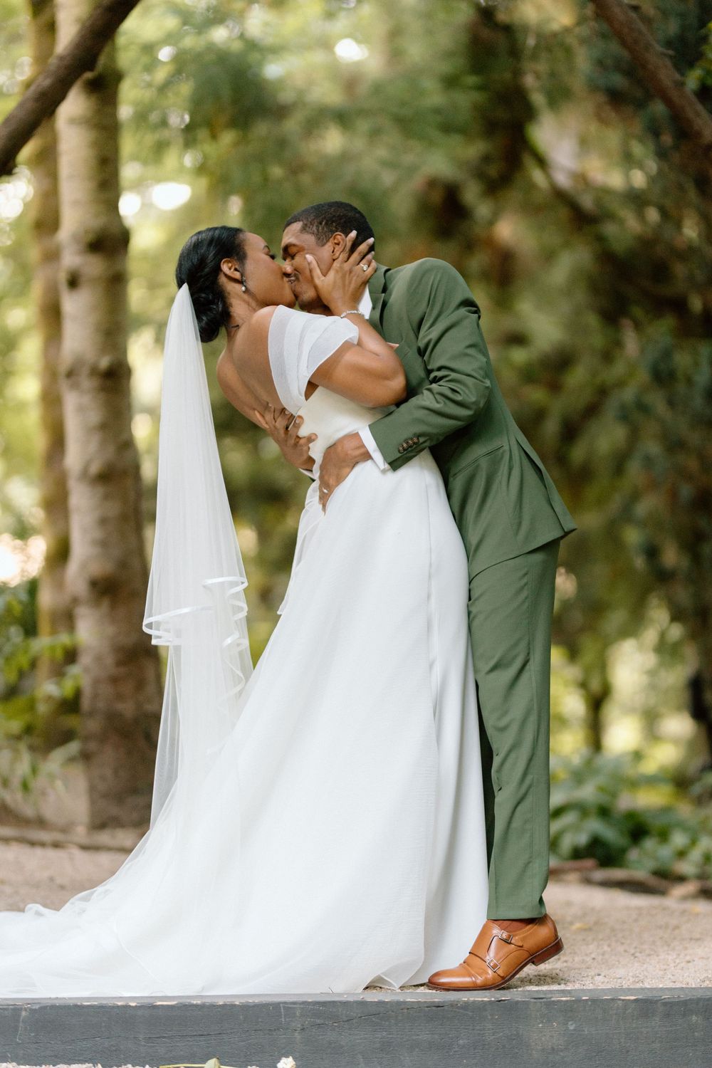 A romantic wedding couple shares a kiss outdoors, with the bride in a flowing white gown and groom in a green suit.