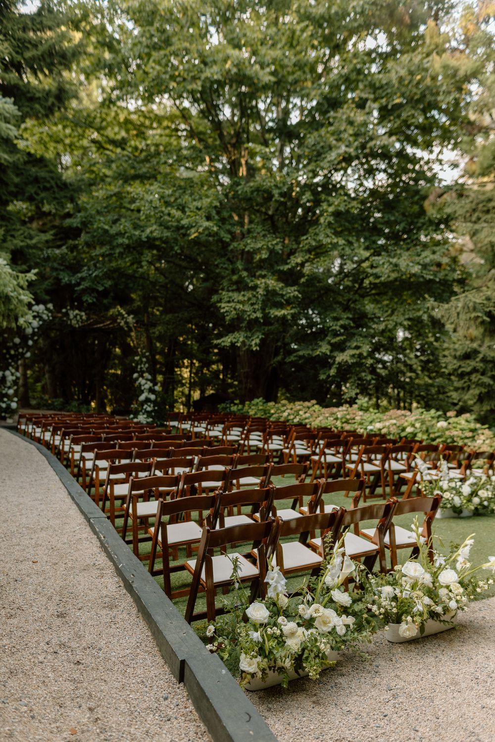 Wooden ceremony chairs arranged in curved rows along a gravel path with lush greenery and white floral decorations.