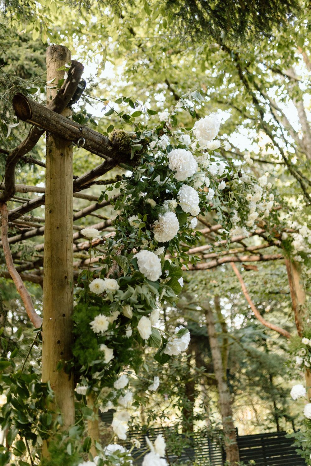 White roses and floral arrangements decorate a rustic wooden pergola in a lush garden setting.