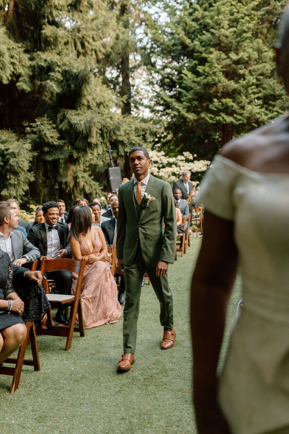 A groomsman in a stylish green suit walks down the aisle at an outdoor wedding ceremony surrounded by seated guests.