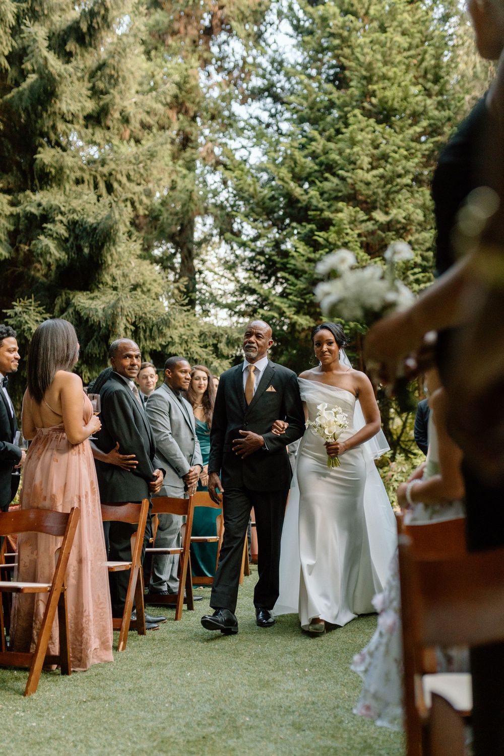 An outdoor wedding ceremony takes place on a green lawn surrounded by tall evergreen trees on a sunny day.