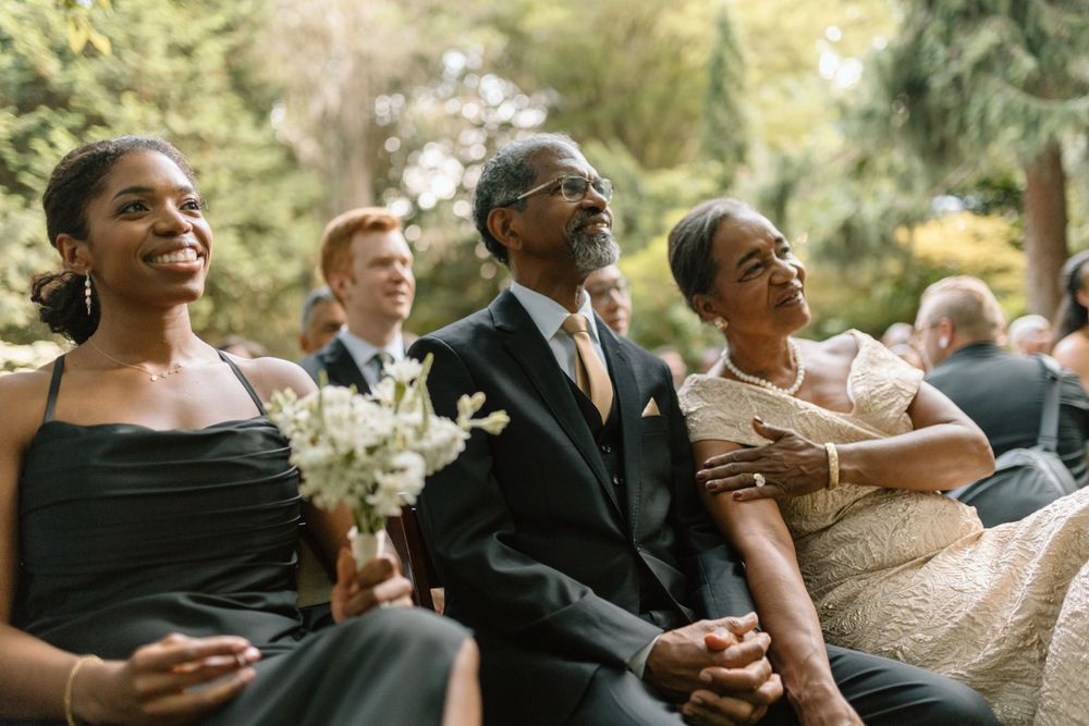 Wedding guests seated in formal attire smile and laugh during an emotional outdoor ceremony moment.