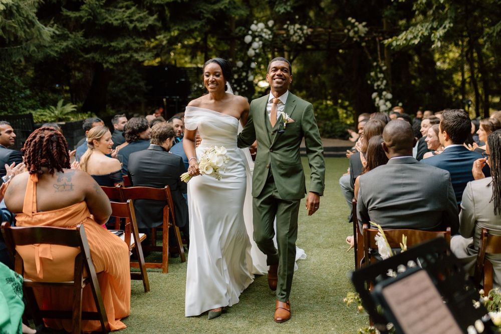 Happy newlyweds walk down the aisle at an outdoor garden wedding ceremony while guests look on from wooden chairs.
