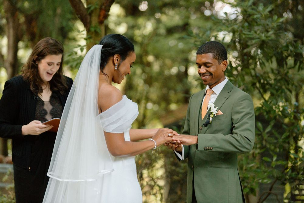An outdoor wedding ceremony takes place in a lush green garden setting as a couple exchanges rings.