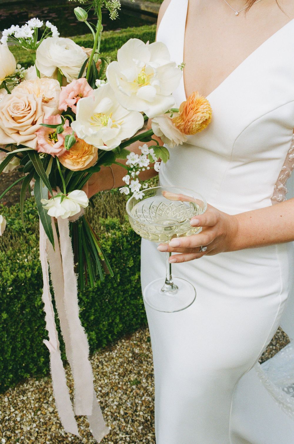 Elegant white wedding dress detail with peach and cream floral bouquet and champagne glass held against outdoor greenery.