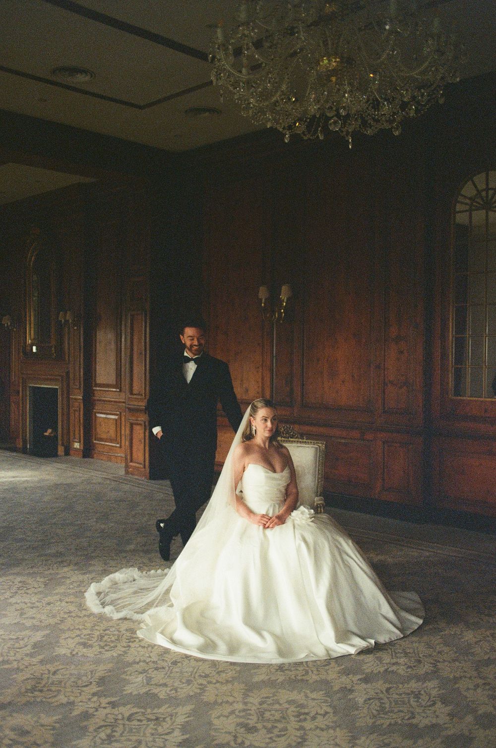 A couple poses in an elegant wood-paneled room with a crystal chandelier for their wedding portrait.