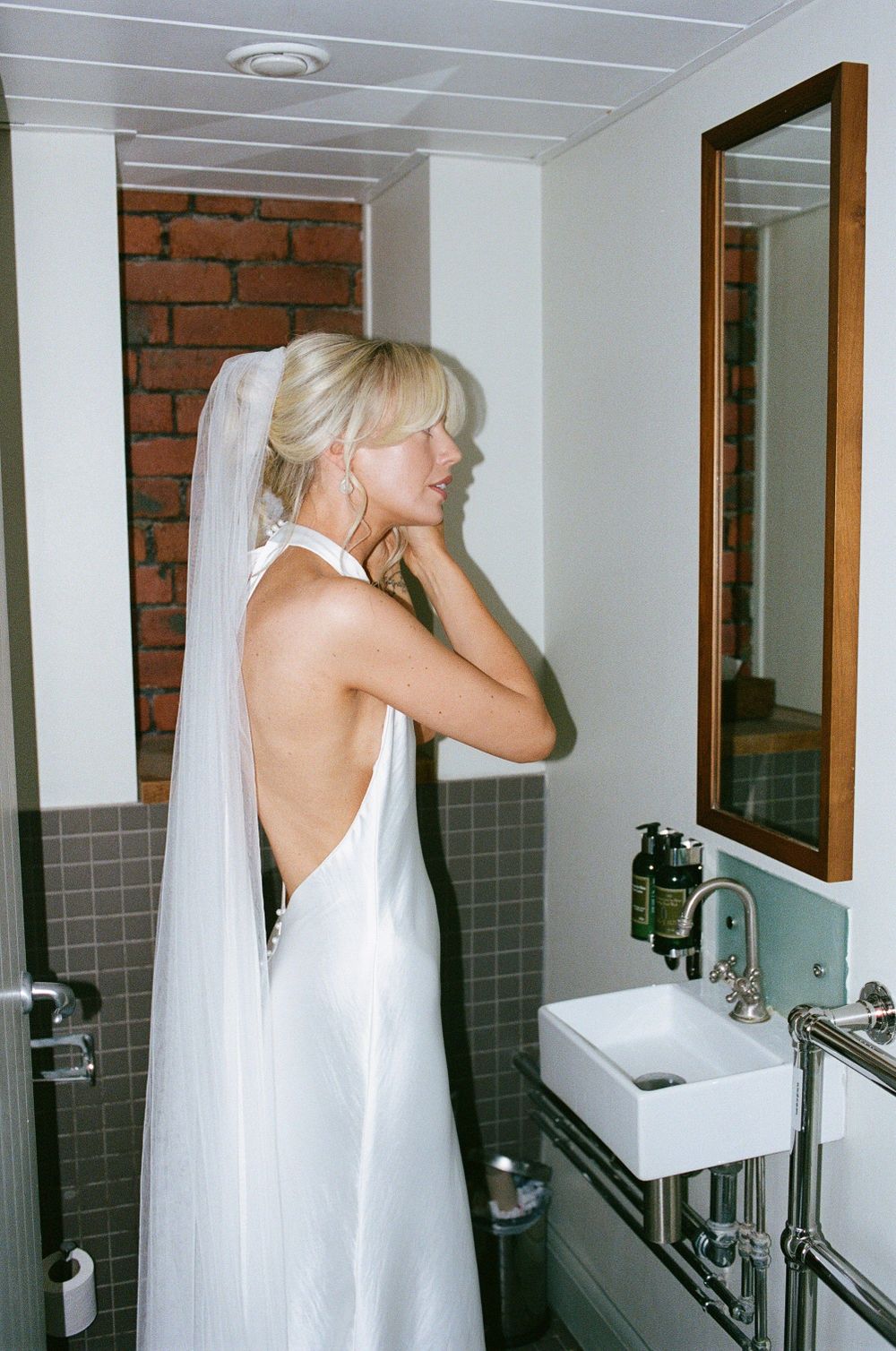 A person in a white halter dress stands in a modern bathroom with exposed brick walls and a framed mirror.