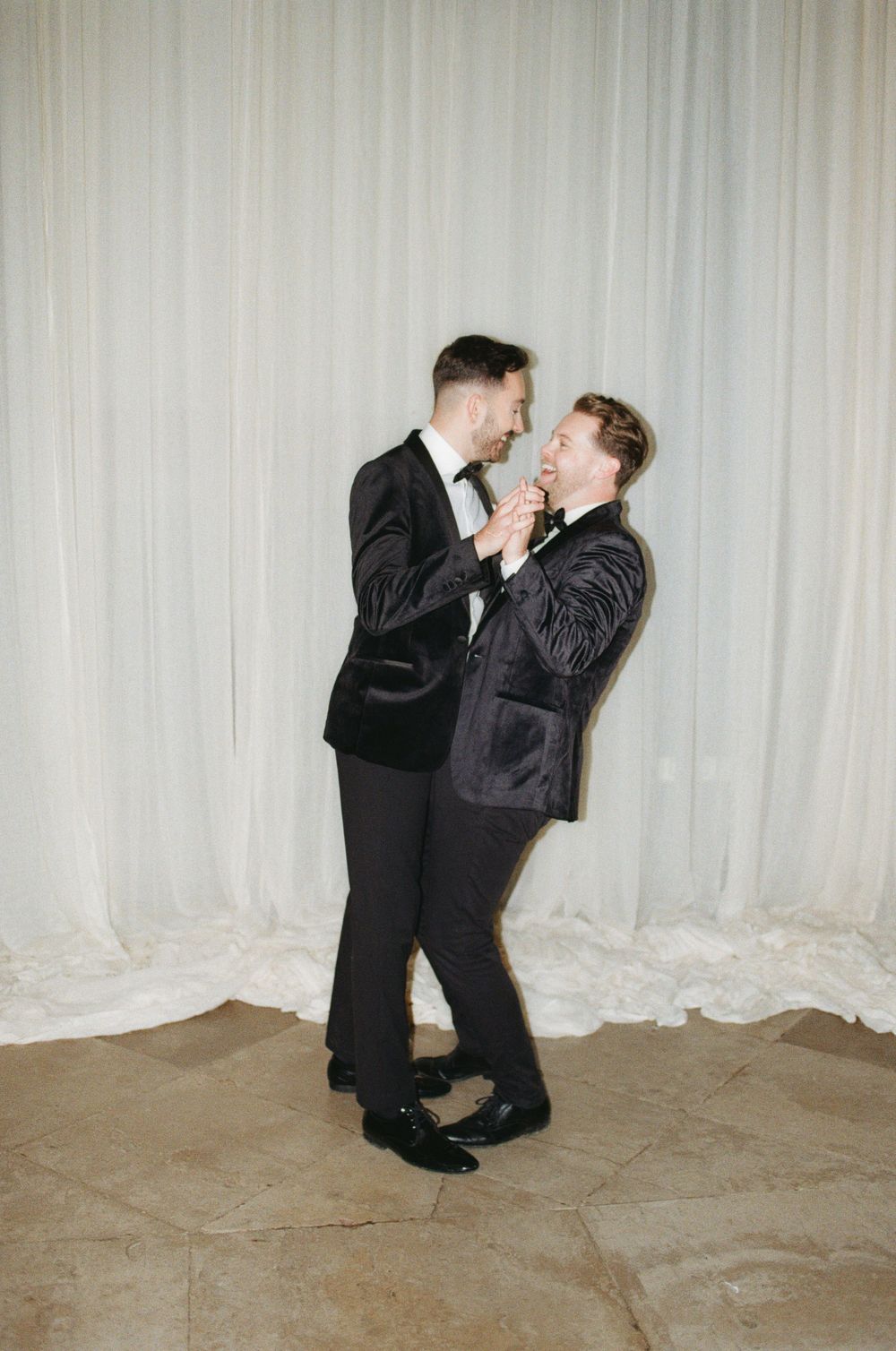 Two people in dark suits share a joyful moment dancing and laughing together against a light curtain backdrop.