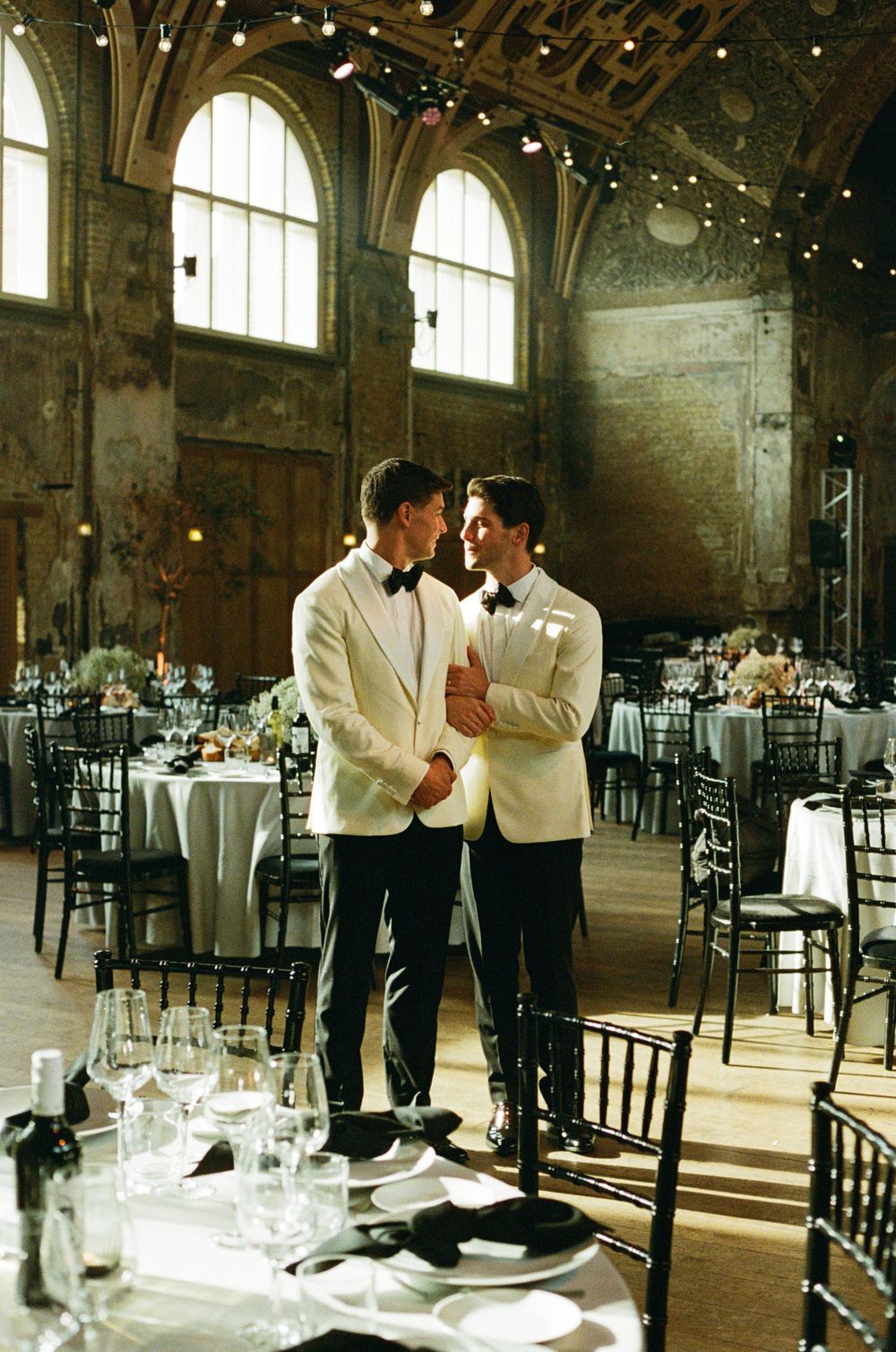 A romantic wedding reception setup in a grand historic venue with vaulted ceilings, arched windows and elegant table settings.