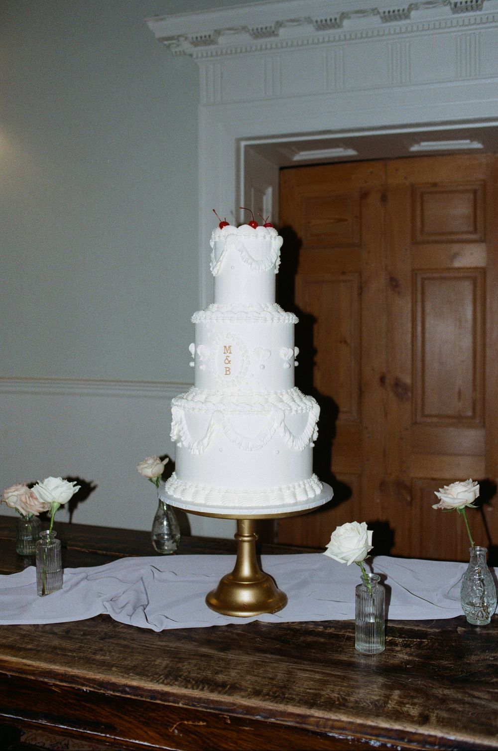 A three-tiered white wedding cake decorated with lace details sits on a gold stand with white roses in glass vases as decor.