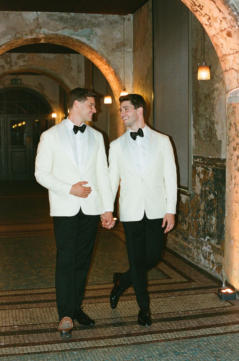 Two people in classic white dinner jackets and bow ties walk together through an arched stone hallway at night.