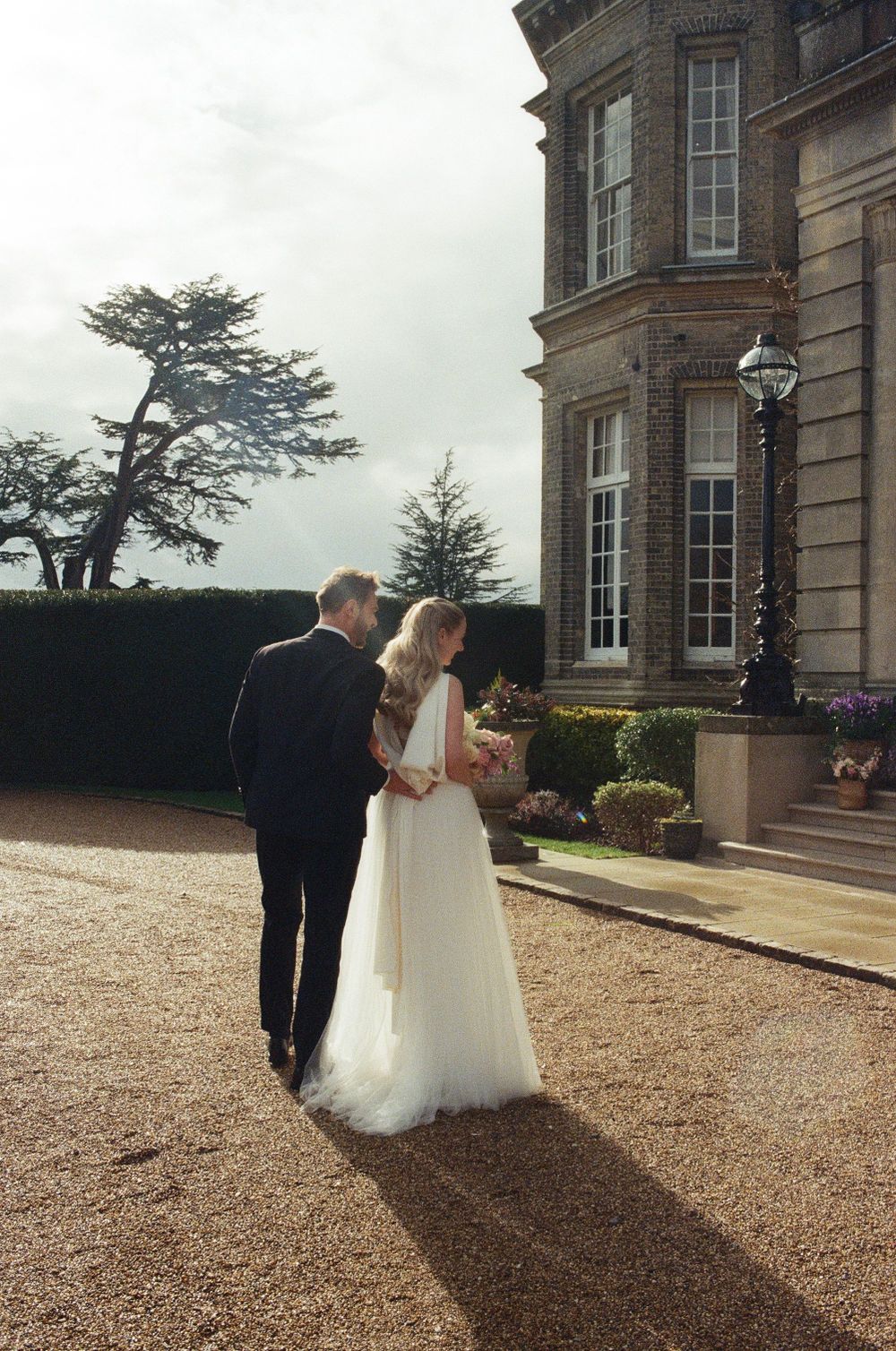 A couple in wedding attire stands together on the gravel driveway of a grand historic mansion on an overcast day.