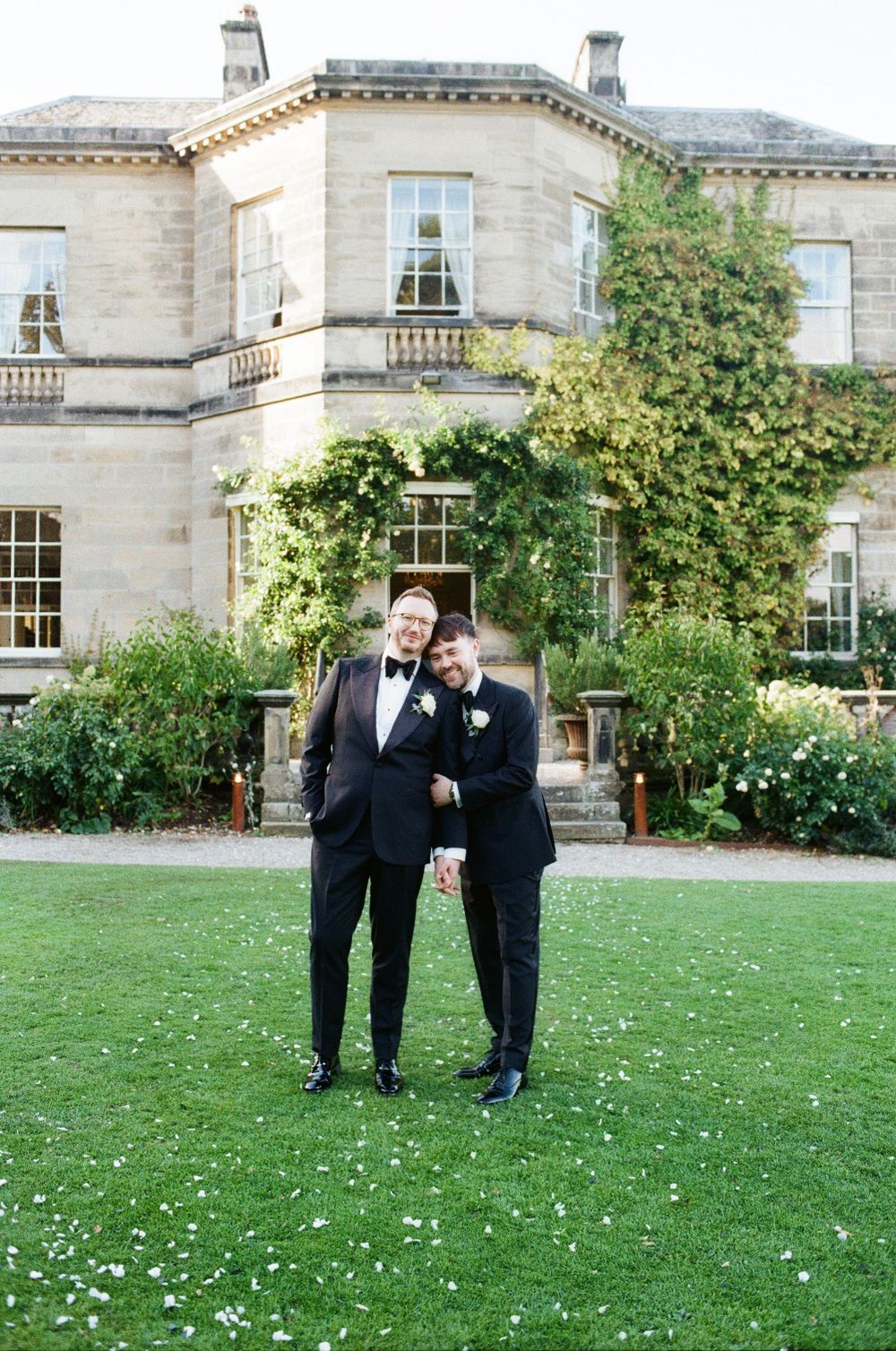 Two people in formal black attire stand on a manicured lawn in front of an elegant stone mansion with climbing vines.