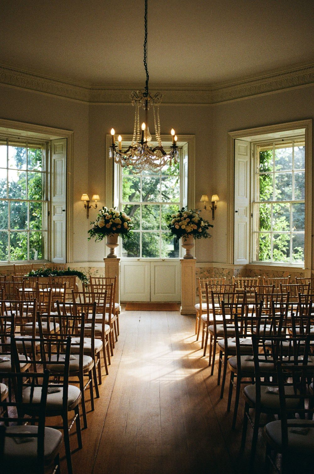 Elegant indoor wedding ceremony setup with wooden chairs, chandeliers, and floral arrangements flanking bright windows.