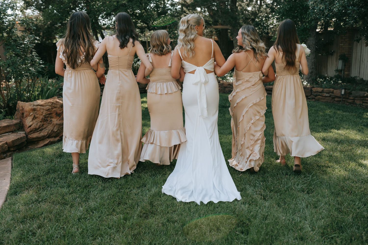Bridesmaids in champagne dresses walk arm in arm with the bride in white across a lawn.