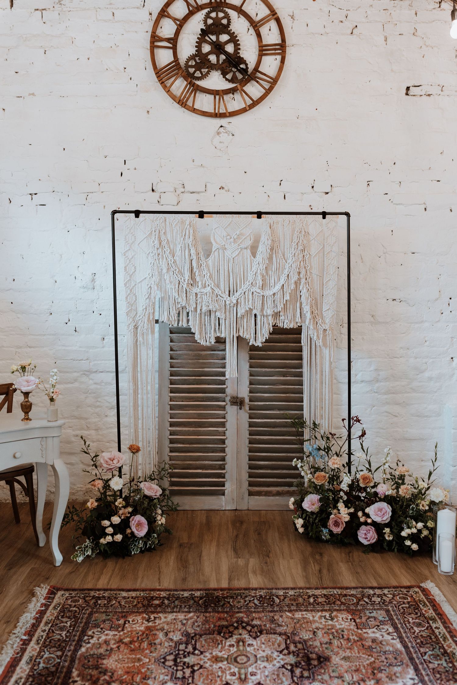 Rustic wedding backdrop with macrame hanging, vintage clock, oriental rug and floral arrangements against white brick wall.