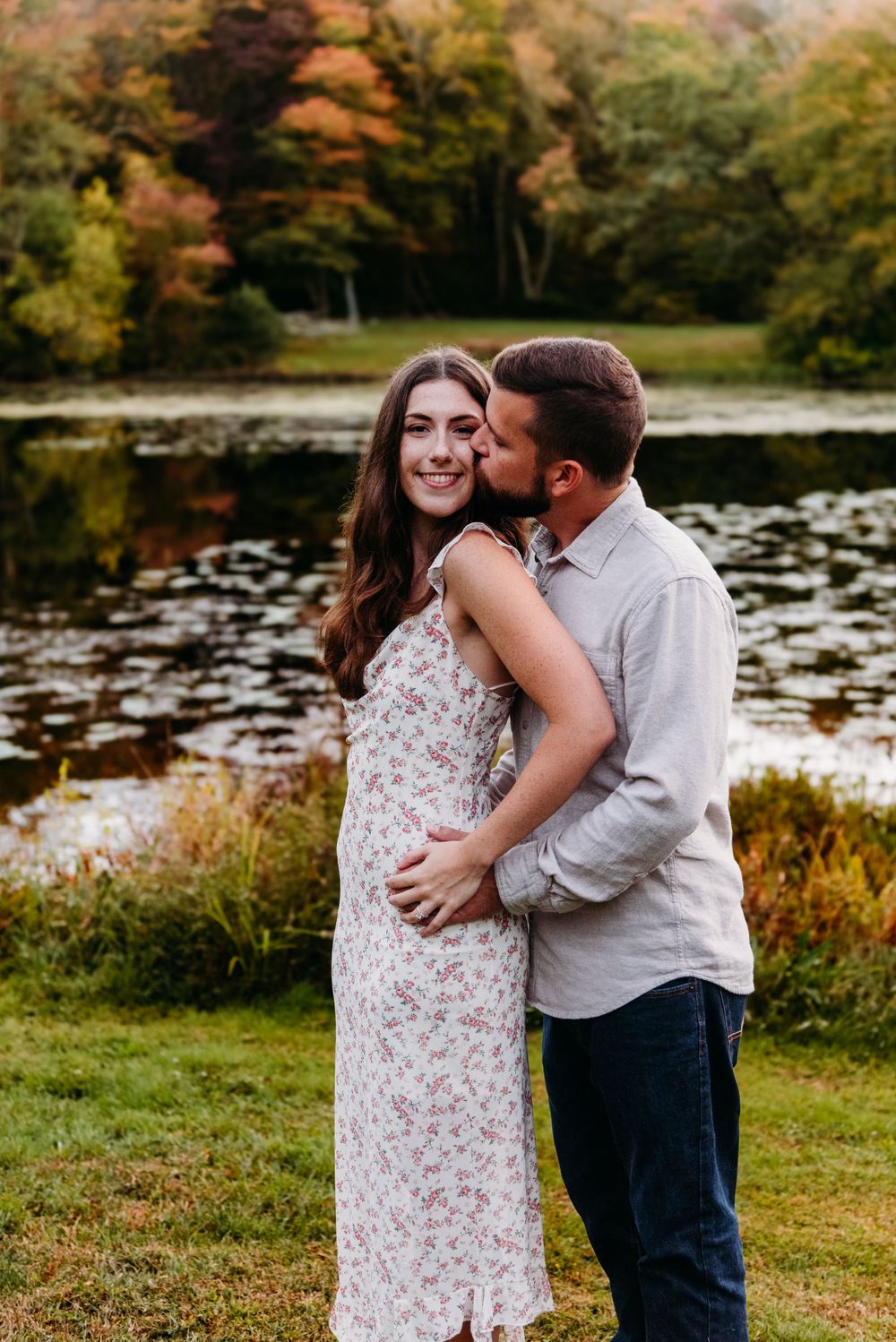 A romantic couple embraces by a serene pond surrounded by autumn foliage in a tranquil outdoor setting.