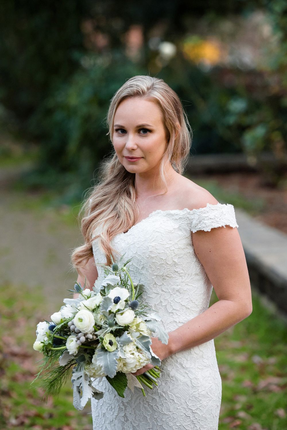 A bride in a lace cap sleeve wedding dress holds a white and green bouquet in an outdoor garden setting.