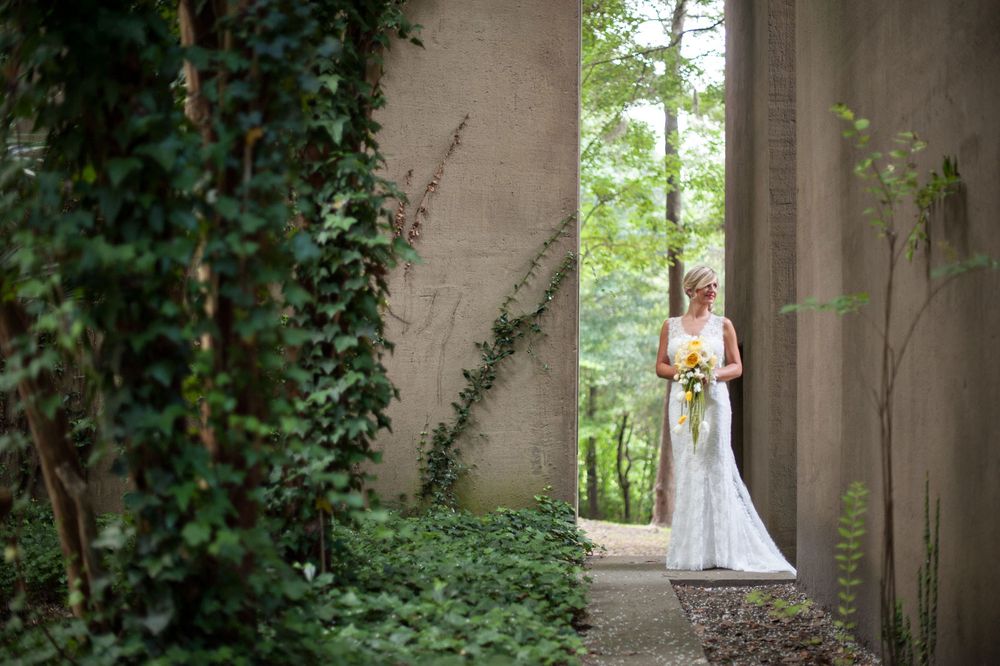 Bride in flowing white gown poses between stone pillars with ivy and greenery in an outdoor garden setting.