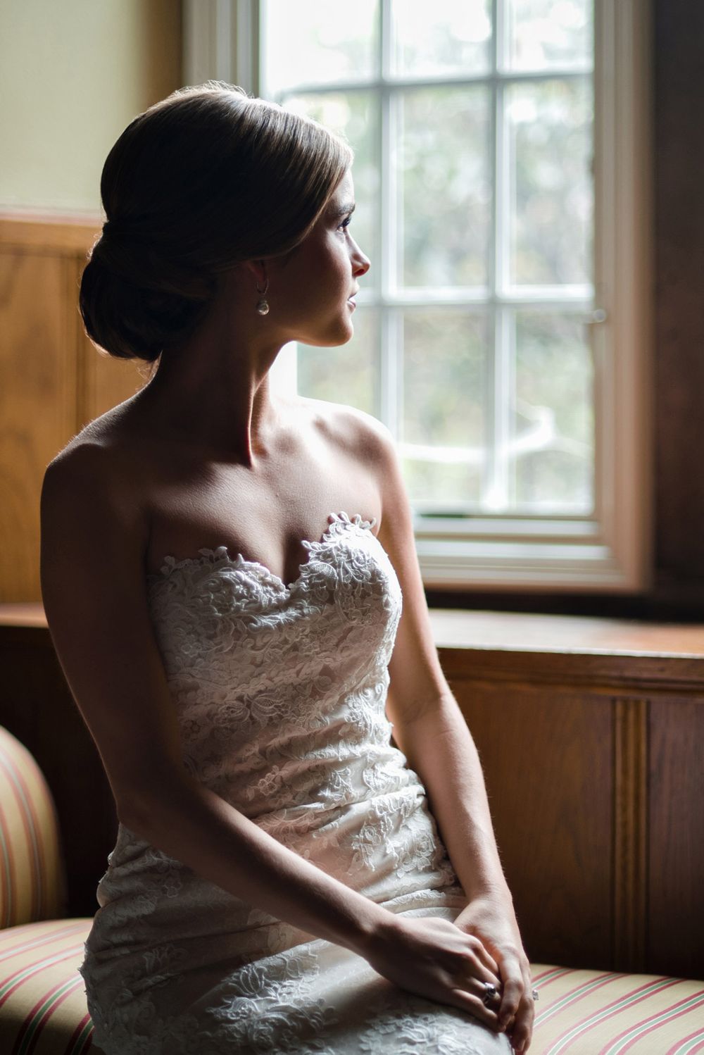 Soft window light illuminates bride in lace dress sitting on vintage furniture.