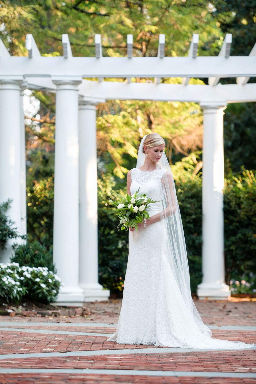 Bride in flowing veil stands beneath white columned pergola holding bridal bouquet.
