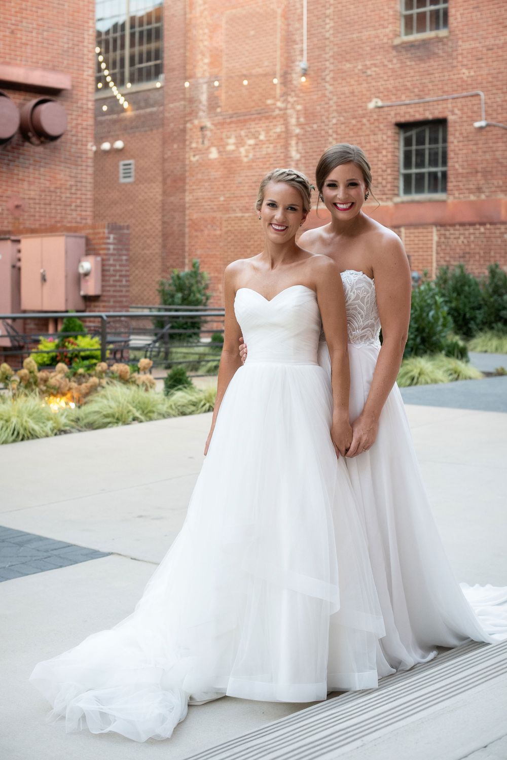 Two brides in elegant white wedding gowns pose together on outdoor terrace with brick building backdrop.