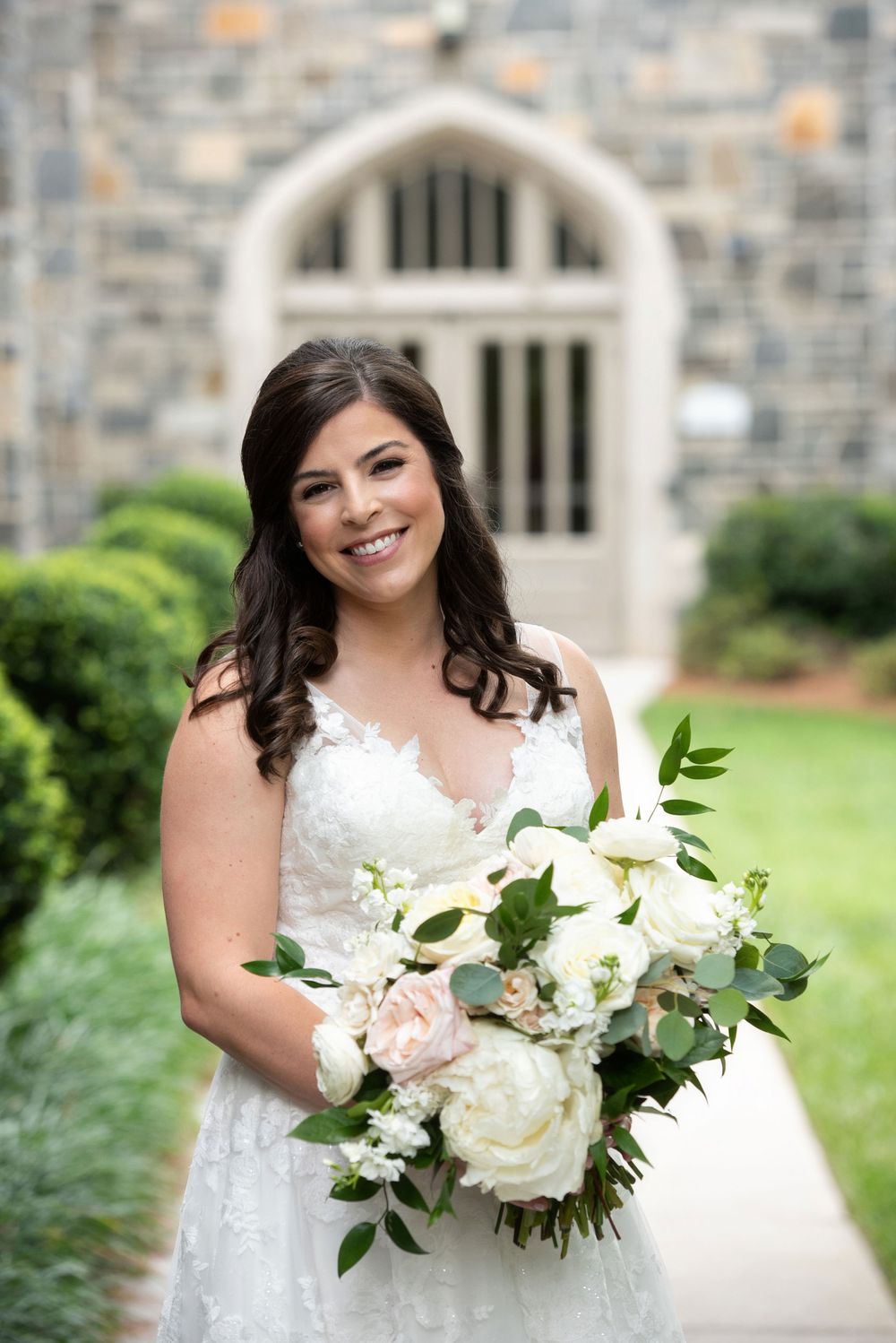 Bride in white lace wedding dress holding elegant bouquet with white peonies and greenery against stone architecture.