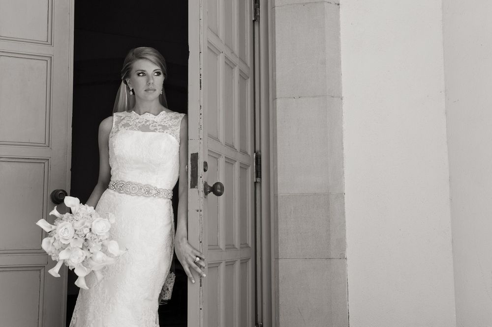 Black and white photo showing a person in an elegant white lace wedding dress with a crystal belt holding a bouquet.