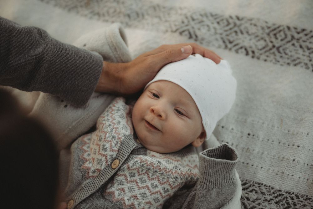 Baby wearing white cap smiles while lying on patterned blanket.